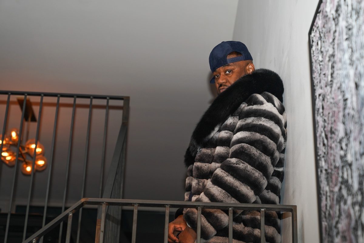 Ghostface Killah in a grey fur coat against a wall above the stair railing