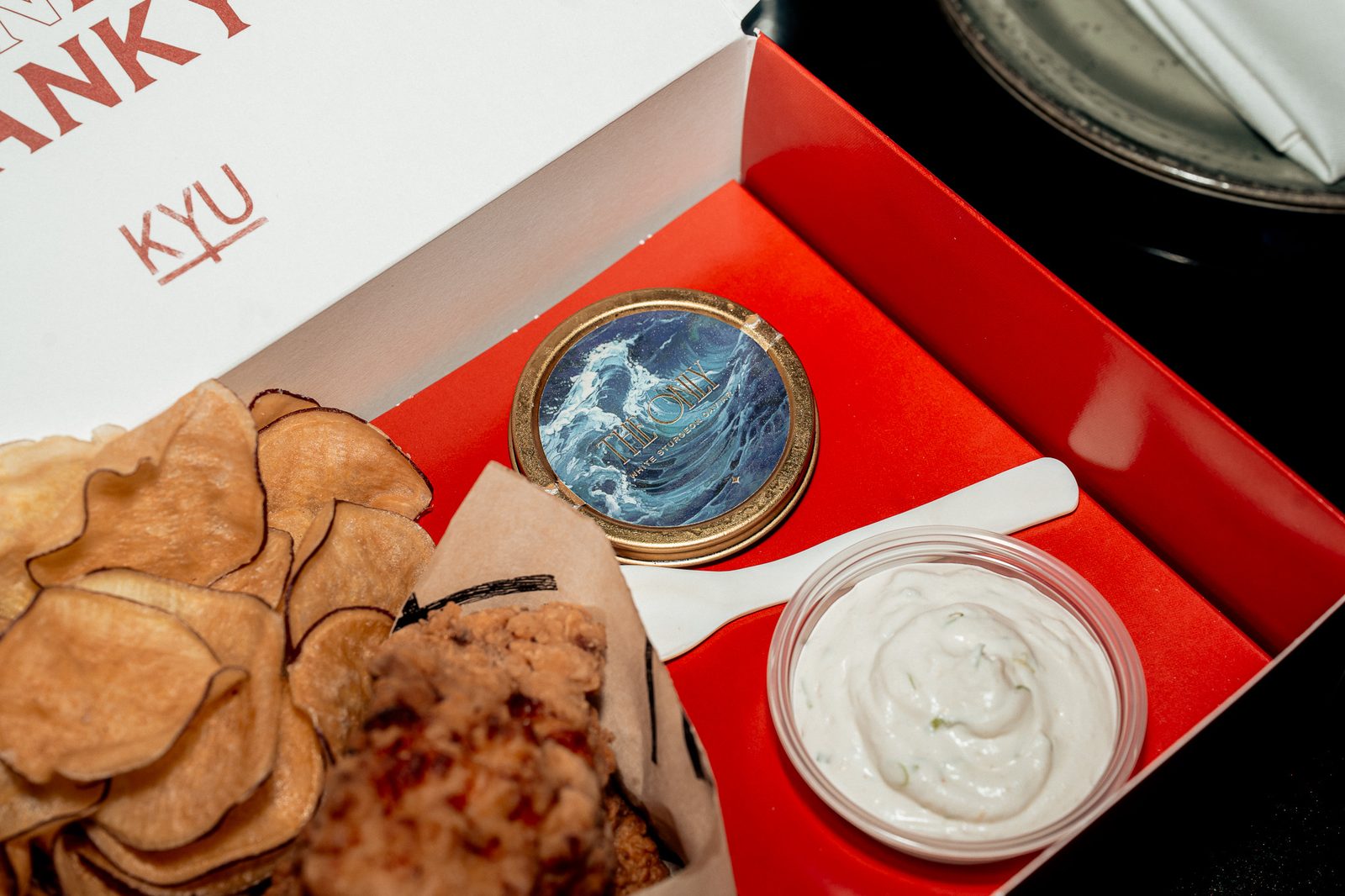 Close-up of the open box: chips, fried chicken, a caviar tin with a wave design and a cup of dip