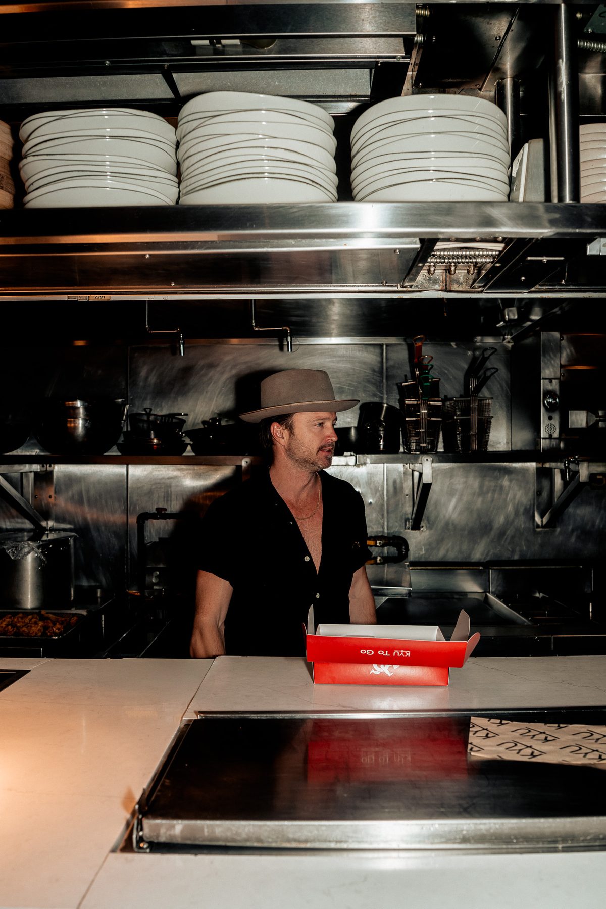 Aaron Paul at the kitchen pass behind stacks of white plates, a red box on the counter
