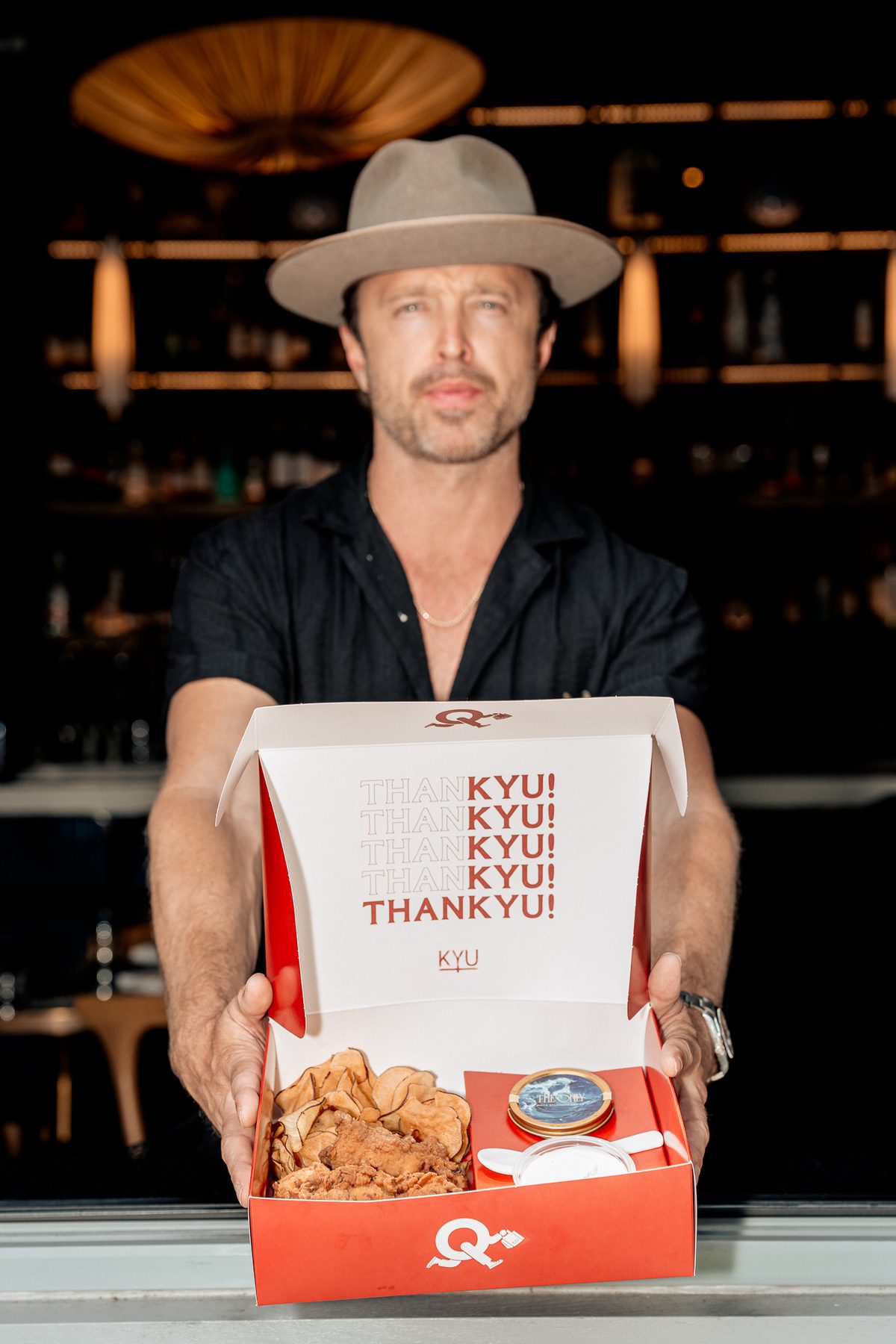 Aaron Paul holding a ThanKYU box out toward the camera under the bar lights