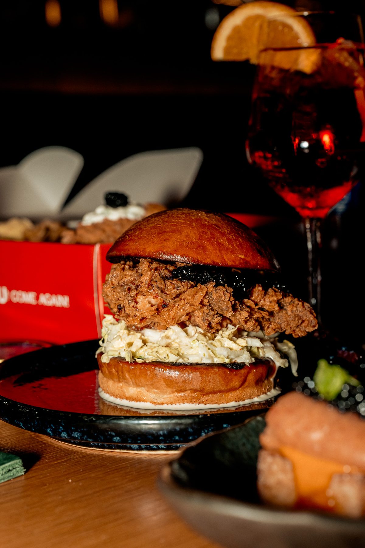A fried chicken sandwich on a dark plate with a cocktail behind it