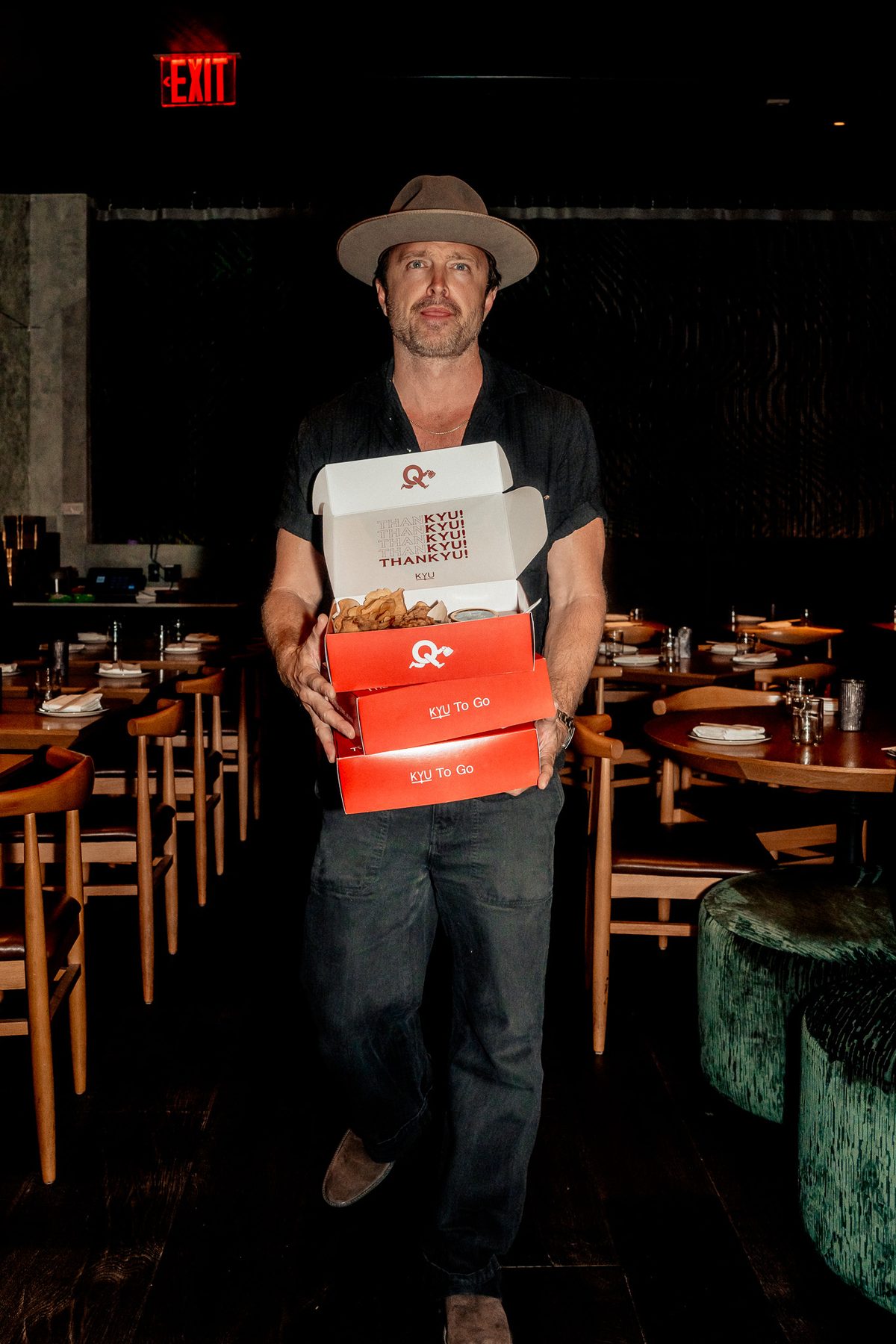 Aaron Paul carrying a stack of ThanKYU boxes through the dining room