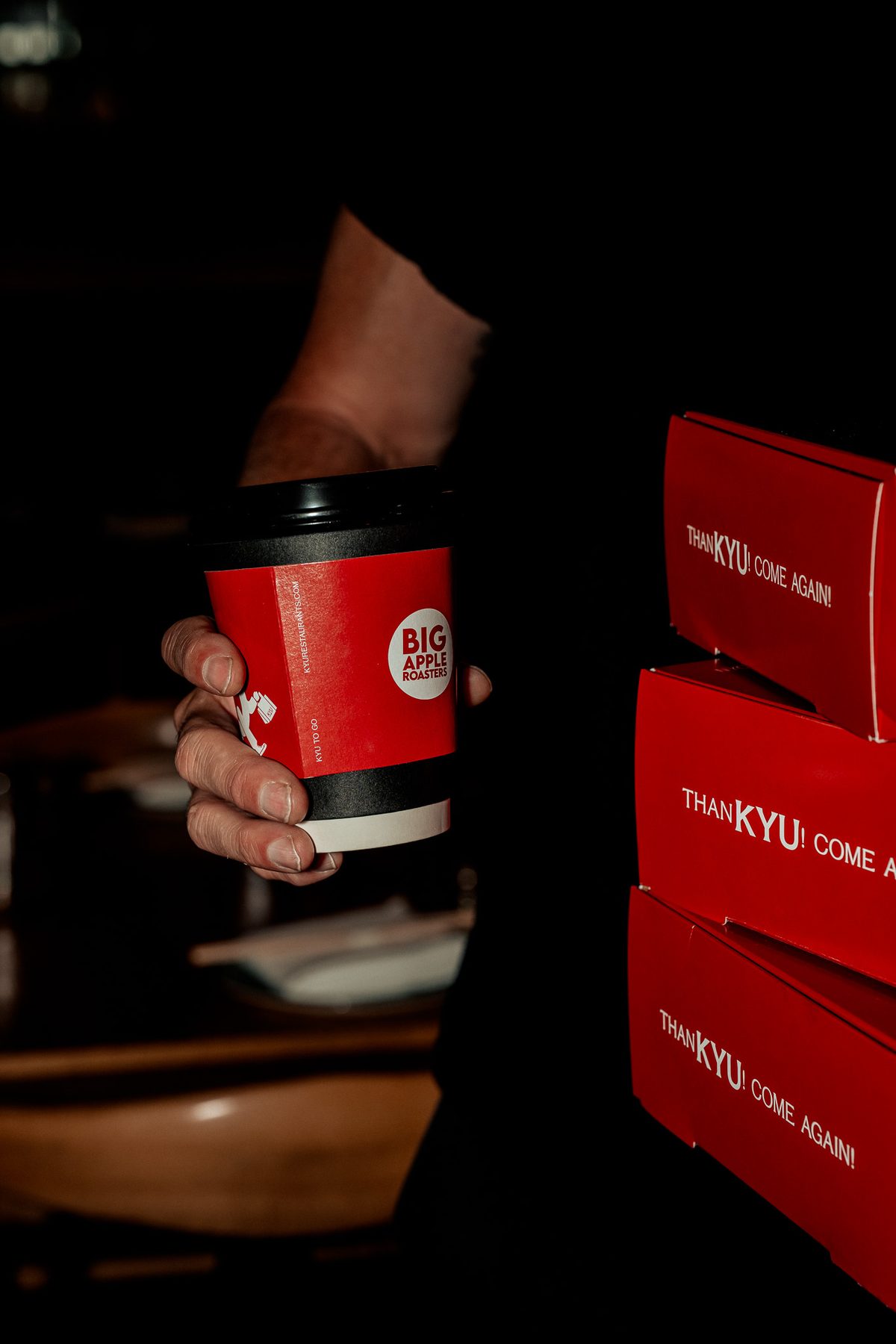 A hand holding a red coffee cup beside a stack of ThanKYU boxes